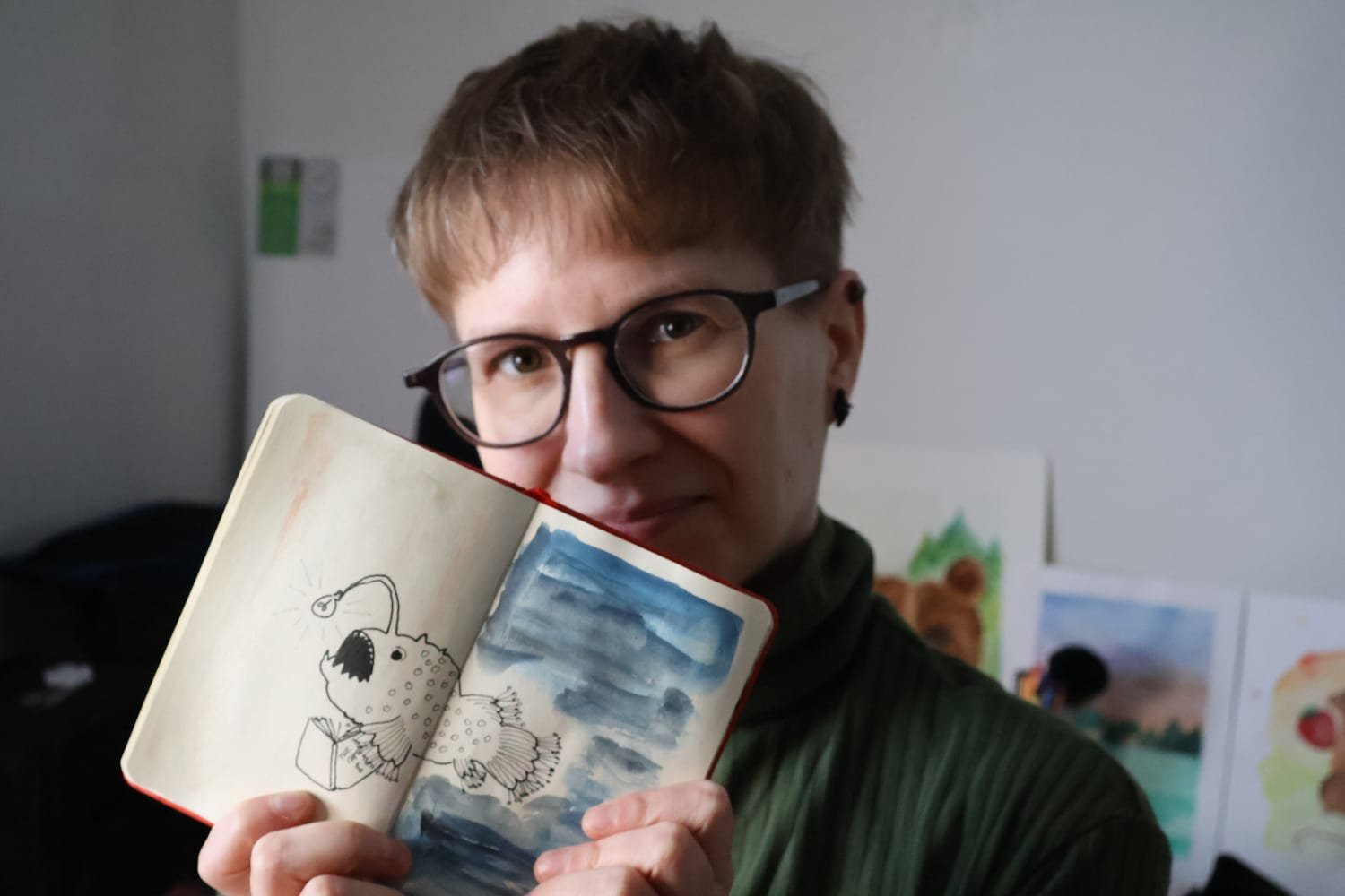 A photo of artist Mervi Eskelinen, a person with short lightish brown hair with longer bangs and shaved sides wearing brown rimmed glasses and a green turtleneck and holding an open sketchbook in front of them with a line drawing of a lampfish with a lightbulb and holding a book on it.