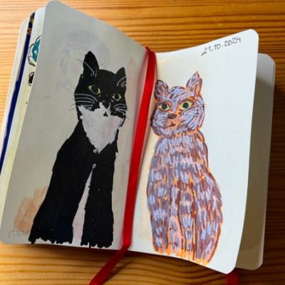 A photo of an open sketchbook on a wooden table with an acrylic marker drawing of a black tuxedo cat and brown gray tabby cat sitting looking straight at you (there's also a Sakura Micron fine liner on the table as well as a metallic clip) by Mervi Eskelinen